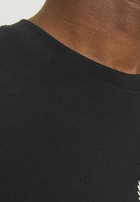 Camiseta de algodón negra con cuello redondo. Textura suave, con un detalle de logo de color claro en el pecho. Vista en primer plano del cuello.
