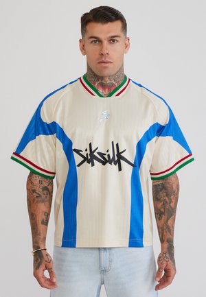 Mand med tatoveringer og næsering iført creme- og blå kortærmet skjorte med rød og grøn kant og sort grafisk tekst foran.