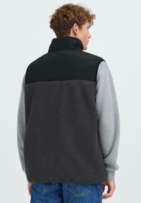 Fleece-Weste in Schwarz und Grau. Sie hat einen hohen Kragen, einen strukturierten Fleece-Körper und ein glattes oberes Panel. Die Rückansicht zeigt eine lockere Passform.