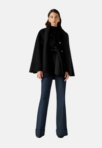 Mantella nera su misura con cintura annodata in vita, abbinata a jeans a zampa blu scuro a vita alta. Tessuto liscio con dettagli hardware minimi.