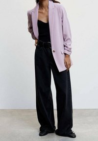 Personne portant un blazer lilas sur un haut noir, avec un pantalon noir à jambes larges, une ceinture noire et des chaussures noires, debout sur un sol en béton.