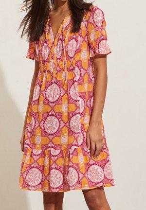 Femme portant une robe courte à manches courtes, longueur genou, avec un motif géométrique et floral orange, rose et blanc, debout devant un fond uni.