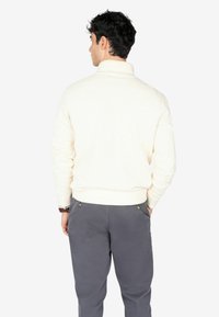 Suéter de beige claro con cuello alto y puños acanalados, combinado con pantalones gris oscuro que cuentan con bolsillos laterales y detalles de botones.