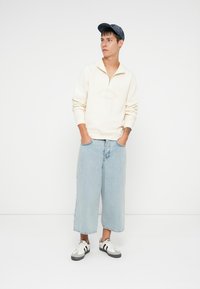 Giovane uomo che indossa un maglione crema a mezza zip, jeans cropped larghi di colore azzurro chiaro, sneakers bianche e un cappellino blu, in piedi con le mani in tasca.