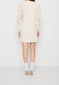 Crèmekleurige jurk met doorschijnende mouwen met bloemenmotief, een afgeronde halslijn en een ontspannen pasvorm, gecombineerd met witte sokken en transparante schoenen met hak.