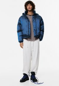 Kurtka puchowa w błyszczącym ciemnoniebieskim kolorze, z kapturem i zatrzaskami. Noszona na pasiastej koszuli, w połączeniu z jasnoszarymi spodniami dresowymi i botkami.