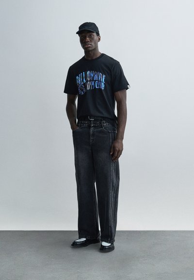 Μαύρο μπλουζάκι με μπλε γραφικό κείμενο, άνετο σκούρο τζιν παντελόνι και μαύρο καπέλο. Τα παπούτσια έχουν μαύρη βάση με λευκές λεπτομέρειες.