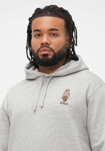 Vīrietis ar bārdu un dreads, ģērbies pelēkā kapučjakā ar Polo lāča logotipu, sēž krustām kājām uz krūtīm.