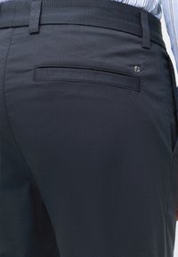 Gros plan sur un pantalon habillé bleu marine foncé avec une unique poche arrière passepoilée et un petit logo métallique près de la poche, porté avec une chemise rayée.
