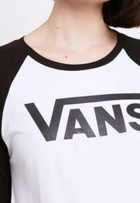 Hvid baseball t-shirt med sorte raglanærmer, der har et stort sort "VANS" logo trykt på forsiden. Blød bomuldsstruktur.