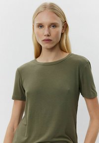 Alyvuogių žalios spalvos glamžyta marškinėliai su trumpomis rankovėmis ir apvalia iškirpte, pasižymintys minkšta tekstūra ir prigludusia siluetu.