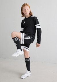 Chemise noire à manches longues avec des rayures blanches, short noir avec des accents blancs, chaussettes noires montantes jusqu'aux genoux et chaussures de football blanches.