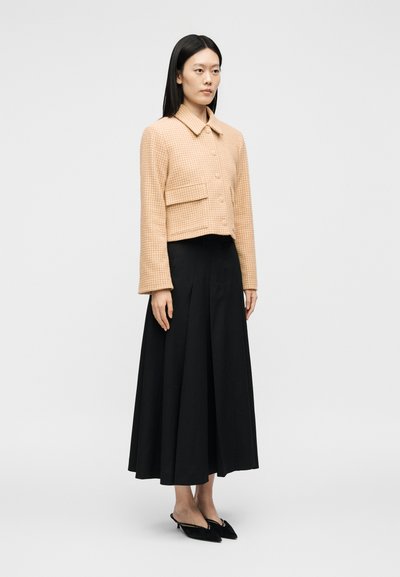 Femme portant une veste courte beige à carreaux avec des boutons, un pantalon noir taille haute à jambes larges et des chaussures slingback noires à bout pointu, debout immobile.