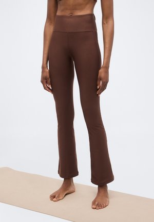 Osoba stojí bosá na béžovej podložke, má na sebe hnedé rozšírené jogové nohavice s vysokým pásom a zodpovedajúci krátky top.