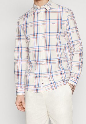 Hombre con camisa blanca de manga larga abotonada con patrón de cuadros azul y rojo y pantalones de color claro, mano en el bolsillo, tatuaje visible.