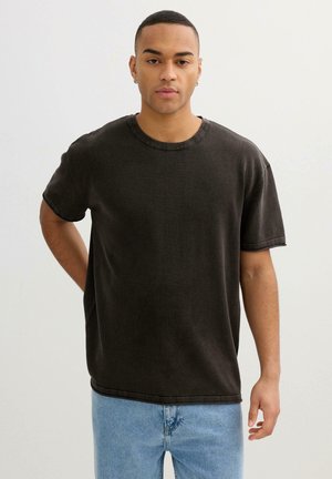 Joven con el pelo corto que lleva una camiseta negra de gran tamaño y unos vaqueros azul claro, de pie frente a un fondo liso y claro.