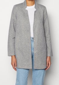 Chaqueta gris tejida con diseño sin cuello, cierre frontal de broches y dos bolsillos delanteros. Llevada sobre una camisa blanca y jeans azul claro.