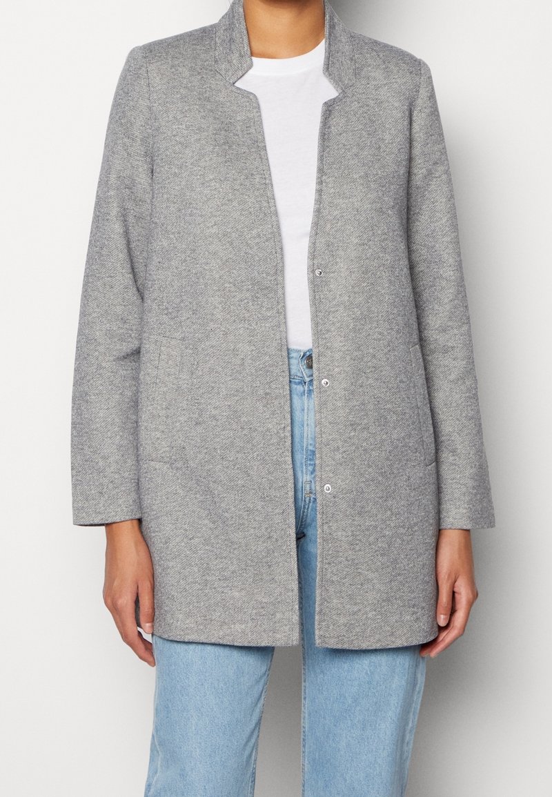 Chaqueta gris tejida con diseño sin cuello, cierre frontal de broches y dos bolsillos delanteros. Llevada sobre una camisa blanca y jeans azul claro.