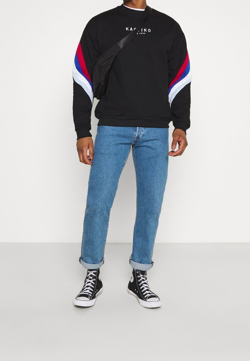 Homme portant un sweat-shirt noir avec des bandes rouges, bleues et blanches sur les manches, un jean bleu retroussé, des baskets montantes noires et un sac bandoulière noir.