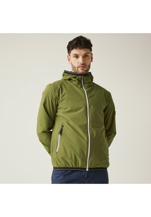 Man draagt een olijfgroene capuchonjas met rits, handen achter de rug, staand tegen een effen lichte achtergrond.