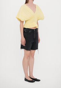 Top giallo a maniche puff con scollo a V profondo e tessuto texturizzato, abbinato a pantaloncini di jeans neri a vita alta con orli sfrangiati e ballerine nere.