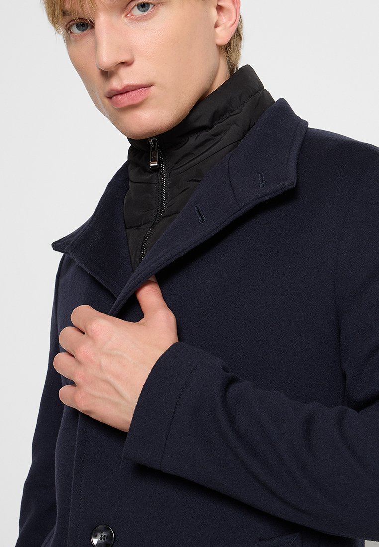 Manteau en mélange de laine marine avec col cranté, fermeture à bouton et doudoune noire zippée visible à l'intérieur, mettant en avant un design superposé.