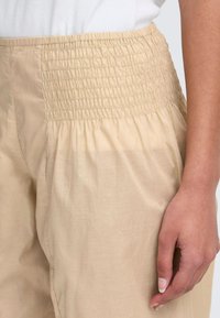 Pantalons beiges avec une taille smockée, présentant une texture lisse et un design ample et fluide. La taille comporte des détails froncés.