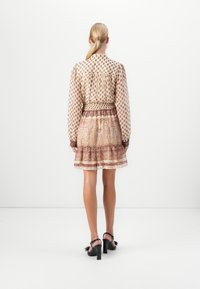 Femme aux cheveux blonds attachés porte un chemisier à manches longues à motifs et une jupe courte assortie avec des sandales noires à talons carrés, debout contre un mur blanc.