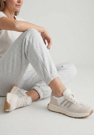 Osoba siedząca na podłodze, ubrana w jasnoszare spodnie dresowe i beżowe sneakersy z białą siateczką i gumowymi podeszwami.