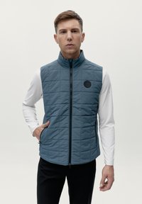Gilet trapuntato senza maniche in tessuto blu chiaro; presenta un colletto alto, chiusura a zip e tasche laterali; accento con logo nero sul petto.