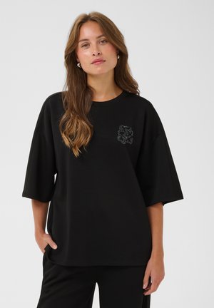 Jeune femme aux longs cheveux portant un top noir oversize à manches courtes avec un petit motif sur la poitrine sur fond clair.