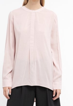Femme portant une blouse rose pâle à manches longues avec un col rond à bouton unique et un pantalon noir ample.