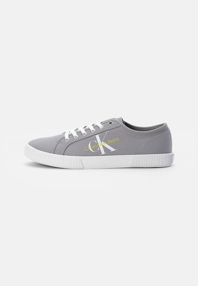 Gray canvas sneakers with white laces and rubber outsole. Features "Calvin Klein Jeans" logo in yellow on the side, subtle texture and design.