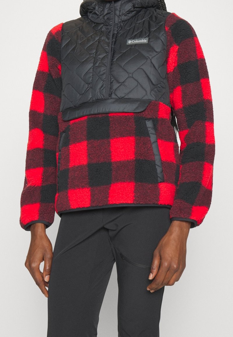 Personne portant une veste polaire rouge et noire à carreaux avec un panneau matelassé noir sur la poitrine et un pantalon noir, debout avec les bras détendus.