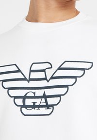 White sweatshirt featuring a navy blue striped eagle logo with “GA” in bold letters, showcasing a smooth texture and crew-neck design.