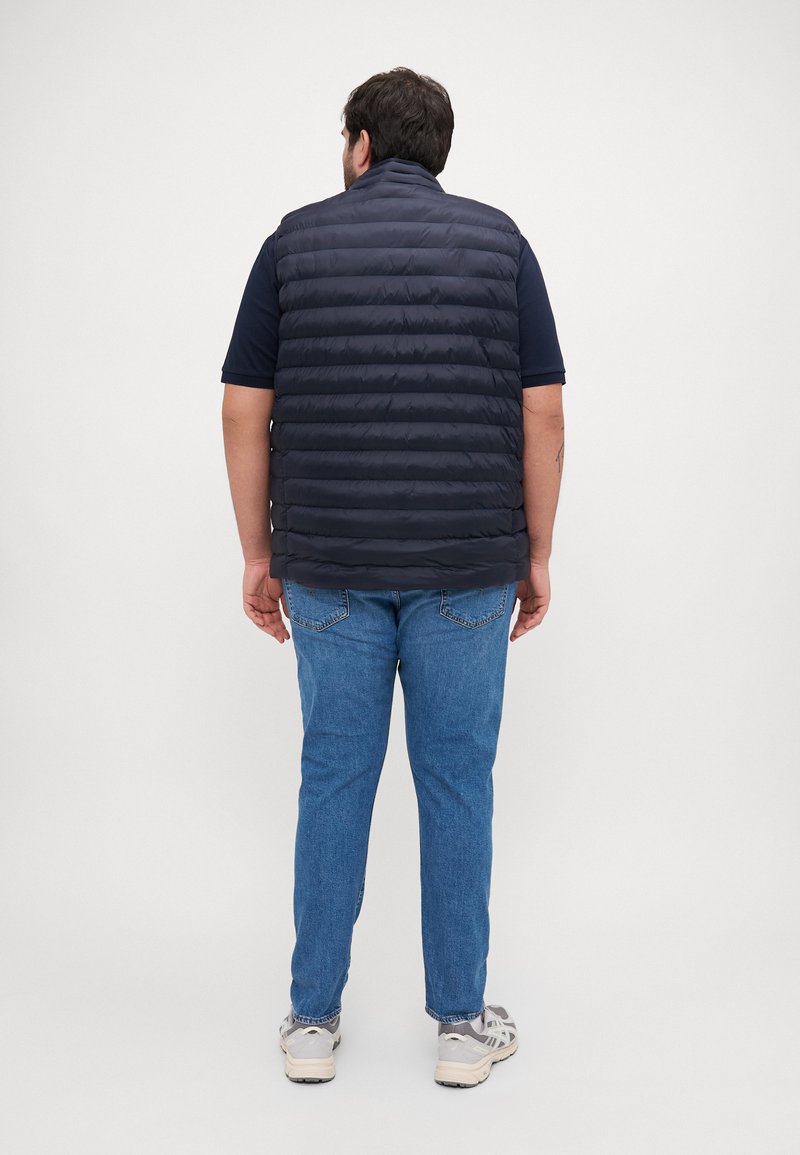 Gilet rembourré bleu marine avec des coutures horizontales, col haut, porté sur une chemise bleu marine, associé à un jean bleu et des baskets de couleur claire.