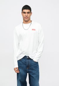 T-shirt de manga longa branca com o logotipo "DIESEL" em vermelho, emparelhada com jeans de denim azul com costura em contraste. O modelo está em pé com as mãos nos bolsos.