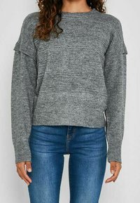 Personne portant un pull en maille grise texturée à manches longues et un jean bleu, debout devant un fond clair uni.