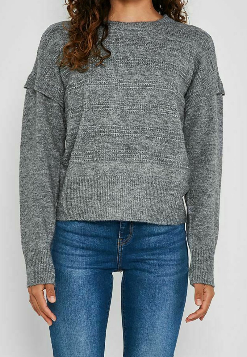 Personne portant un pull en maille grise texturée à manches longues et un jean bleu, debout devant un fond clair uni.