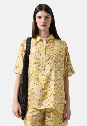 Kobieta ubrana w żółtą koszulę w krótkim rękawie w wzory oraz pasujące spodnie, niosąca dużą czarną plecioną torebkę na ramię na jednolitym tle.