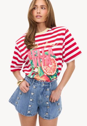 Femme portant un t-shirt rayé rouge et blanc avec le texte "Bella Vita" et un motif de fruits, associé à un short en jean taille haute avec plusieurs boutons.
