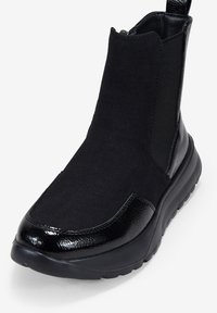 Bottines noires à talon, alliant suede mat et cuir verni brillant. Panneaux élastiques sur les côtés et une languette à l'arrière pour un enfilage facile.