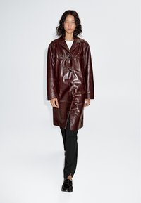 Bordeauxrood leren jas met knoopsluiting aan de voorkant, voorzien van een kraag en een licht oversized ontwerp. Gecombineerd met zwarte, op maat gemaakte broek.