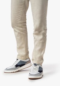 Sneaker bianchi e blu marino con dettagli grigi, caratterizzati da una tomaia texturizzata, design con lacci e una suola in gomma spessa e zigzagata, abbinati a pantaloni slim beige.