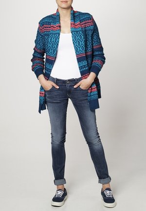 Cardigan tricoté bleu avec des motifs roses et turquoise, superposé à un top blanc. Associé à un jean bleu et des baskets noires.