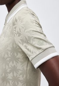 Záber zblízka osoby v svetlo béžovej polo košeli s textúrovanými motívmi palmy a malým okrúhlym logom na rukáve.