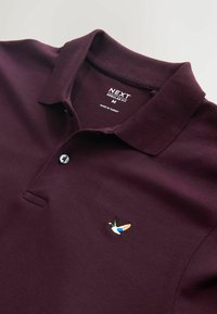 Tricou polo burgundy cu guler clasic, prezintă un emblemă cu rață brodată pe piept și are o guler cu două nasturi. Croială regulară.