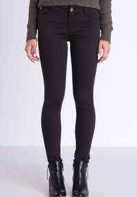 Jeans skinny negros con una textura suave, cintura media y bolsillos frontales. Llevados con botines oscuros ajustados hasta el tobillo.