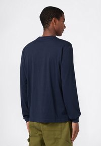 Junge Person mit kurzem Haar, die ein navyblaues Langarmshirt und olivgrüne Hosen trägt, steht seitlich vor einem schlichten weißen Hintergrund.