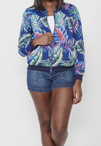 Femme portant une veste bleue à motif floral avec fermeture éclair et un short en denim foncé, tenant la fermeture éclair de la veste d'une main.